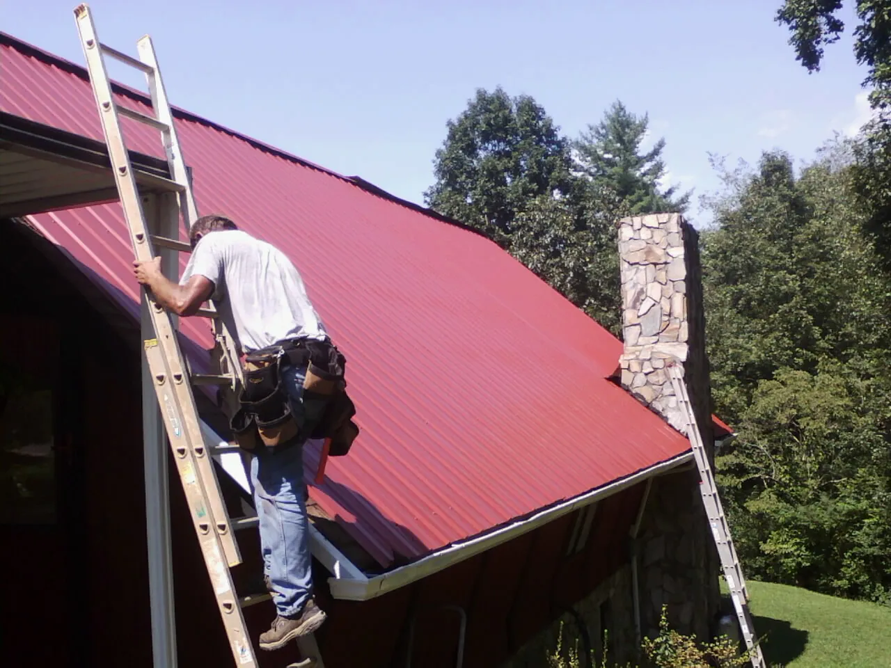 Roofer on a ladder beside a red metal roof and stone chimney — Soffit & Fascia Repair by Roofing Brothers in Monponsett